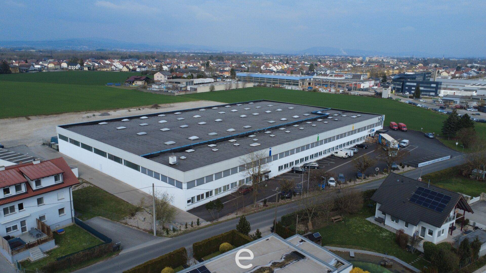 Luftaufnahme eines großen Gewerbegebäudes mit Parkplätzen und umliegender Landschaft.