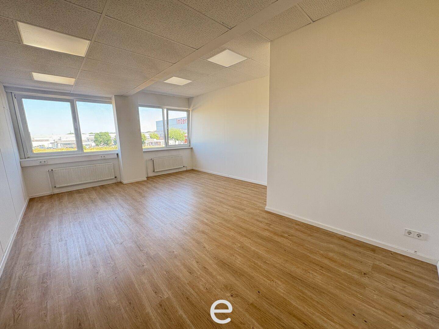 Leeres Büro mit großen Fenstern, Heizkörpern und hellem Laminatboden.