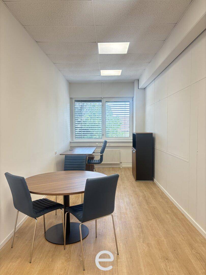 Langes, schmales Büro mit Holzboden, einem runden Tisch und zwei Arbeitsplätzen am Fenster.