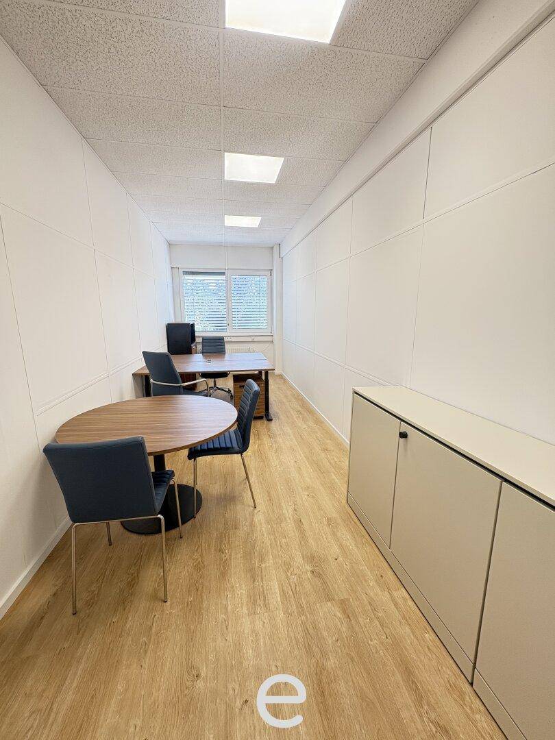 Langes, schmales Büro mit Holzboden, einem runden Tisch und zwei Arbeitsplätzen am Fenster.