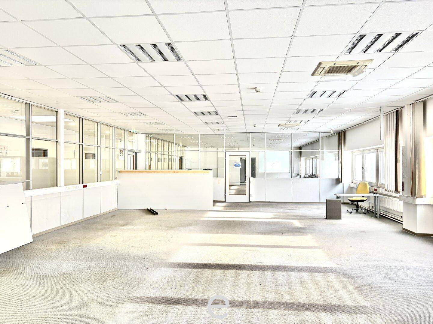 Großes, helles Büro mit Teppichboden, Rasterdecke und modernen Glasabtrennungen für flexible Arbeitsbereiche.