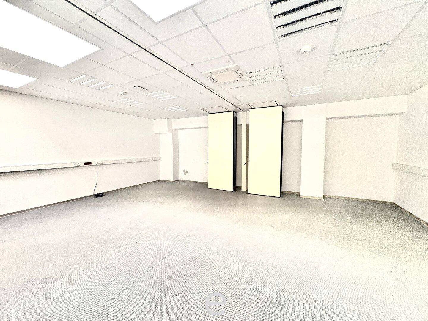 Leerer Büroraum mit hellem Teppichboden, weißen Wänden und einer abgehängten Decke, bereit zur Einrichtung.