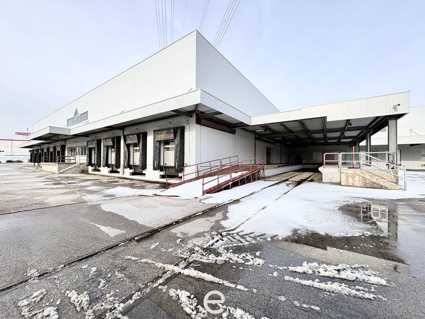 Seitenansicht eines Industriegebäudes mit mehreren Laderampen und einer Zufahrtsrampe, teilweise mit Schnee bedeckt.