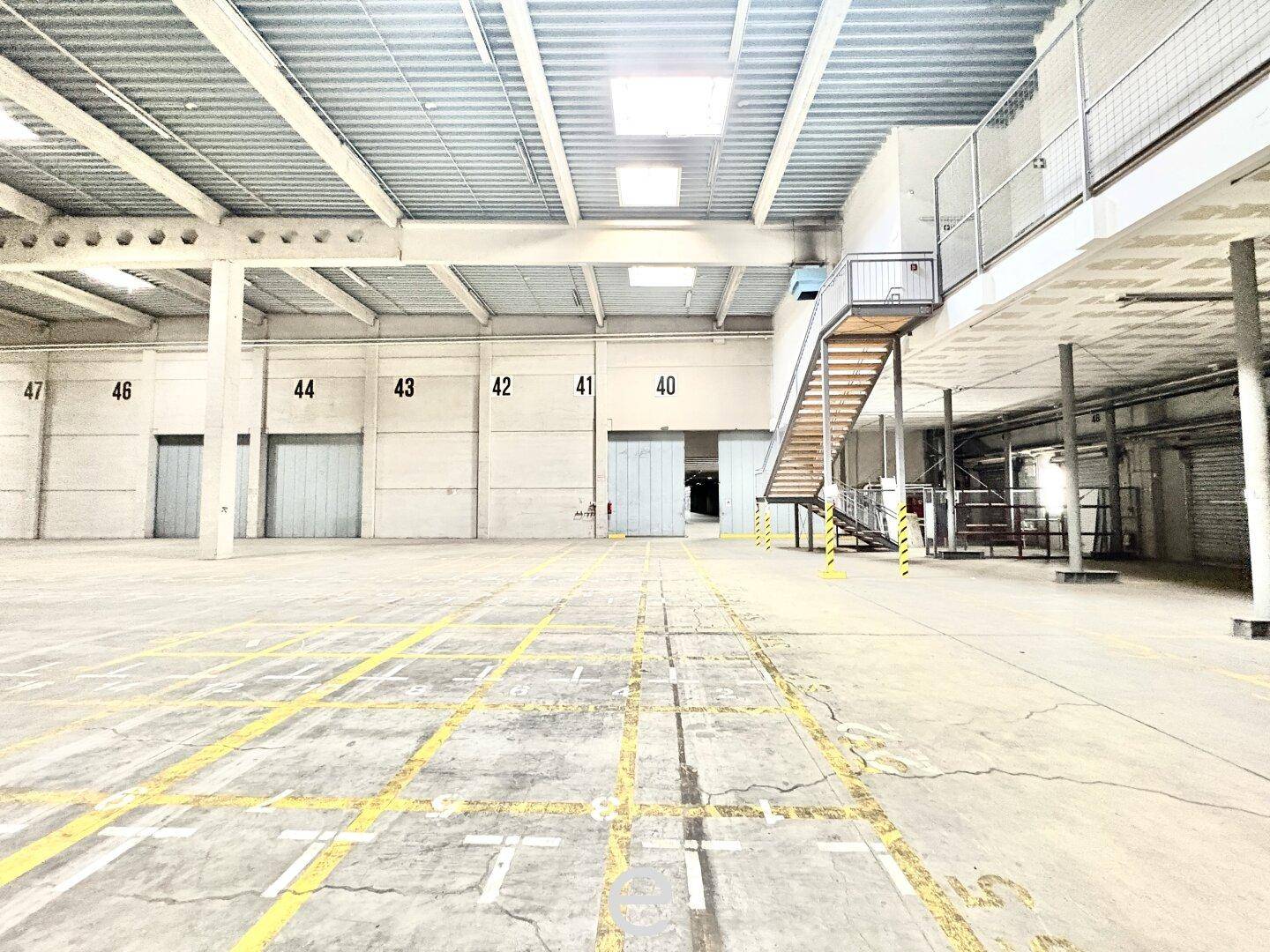 Große, leere Lagerhalle mit Betonboden, gelben Markierungen und einer Metalltreppe, die zu einer oberen Ebene führt.