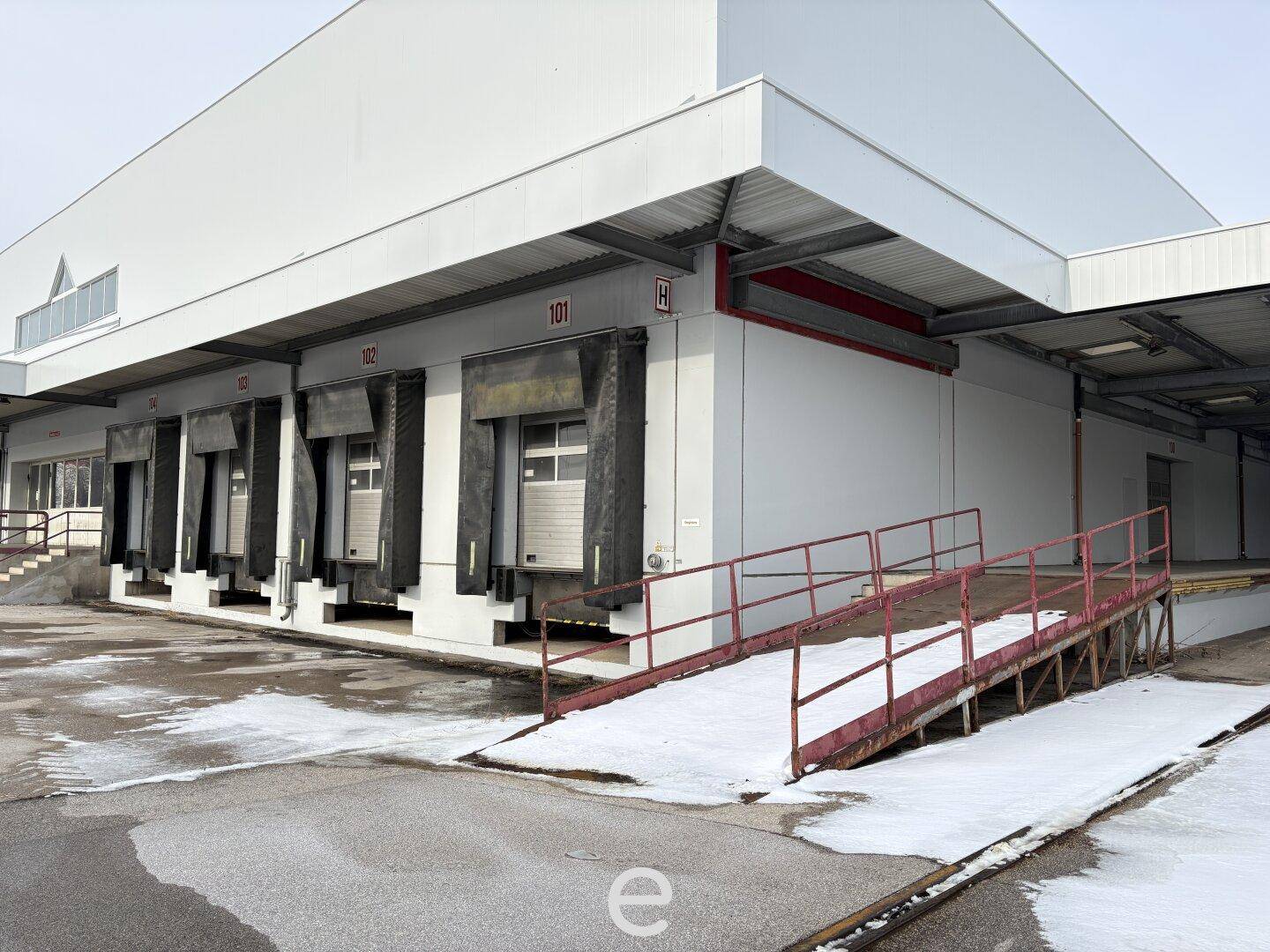 Nahaufnahme der Laderampen und einer roten Zufahrtsrampe an der Fassade eines großen Logistikgebäudes, mit Schnee auf dem Boden.