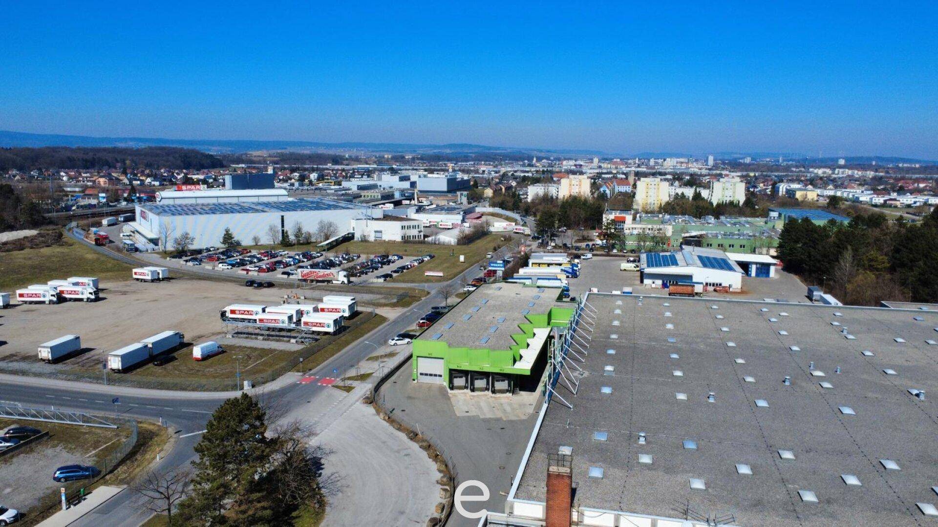 Weitläufige Luftaufnahme eines Gewerbegebiets mit verschiedenen Gebäuden, Parkplätzen und umliegender Infrastruktur.