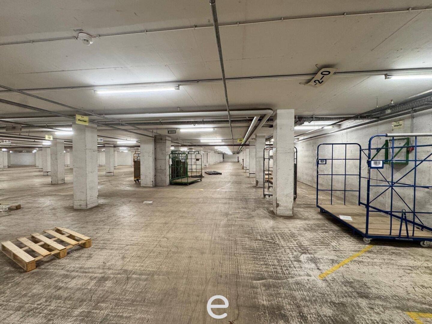 Großer, leerer Lagerraum mit Betonboden, vielen Säulen und einigen Transportwagen, gut beleuchtet.