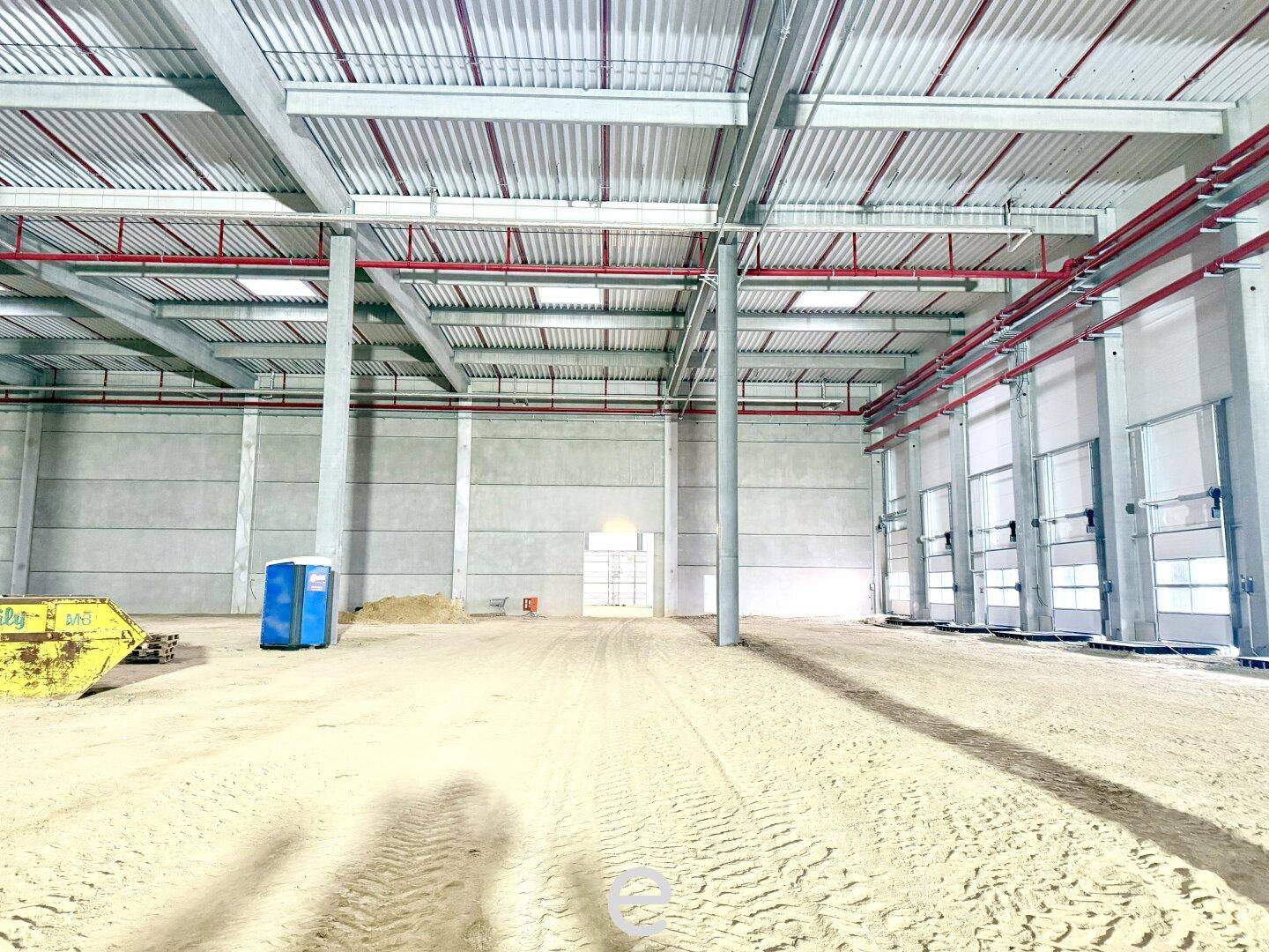 Ein weitläufiger Lagerraum im Bau mit hohen Decken, Betonwänden und sichtbaren roten Leitungen an der Decke.
