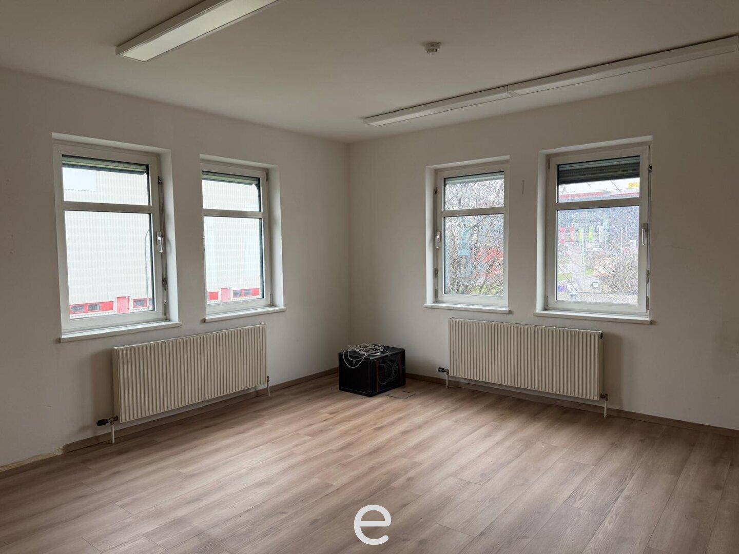 Helles, leeres Büro mit Laminatboden, mehreren Fenstern und Heizkörpern, bereit zur Einrichtung.