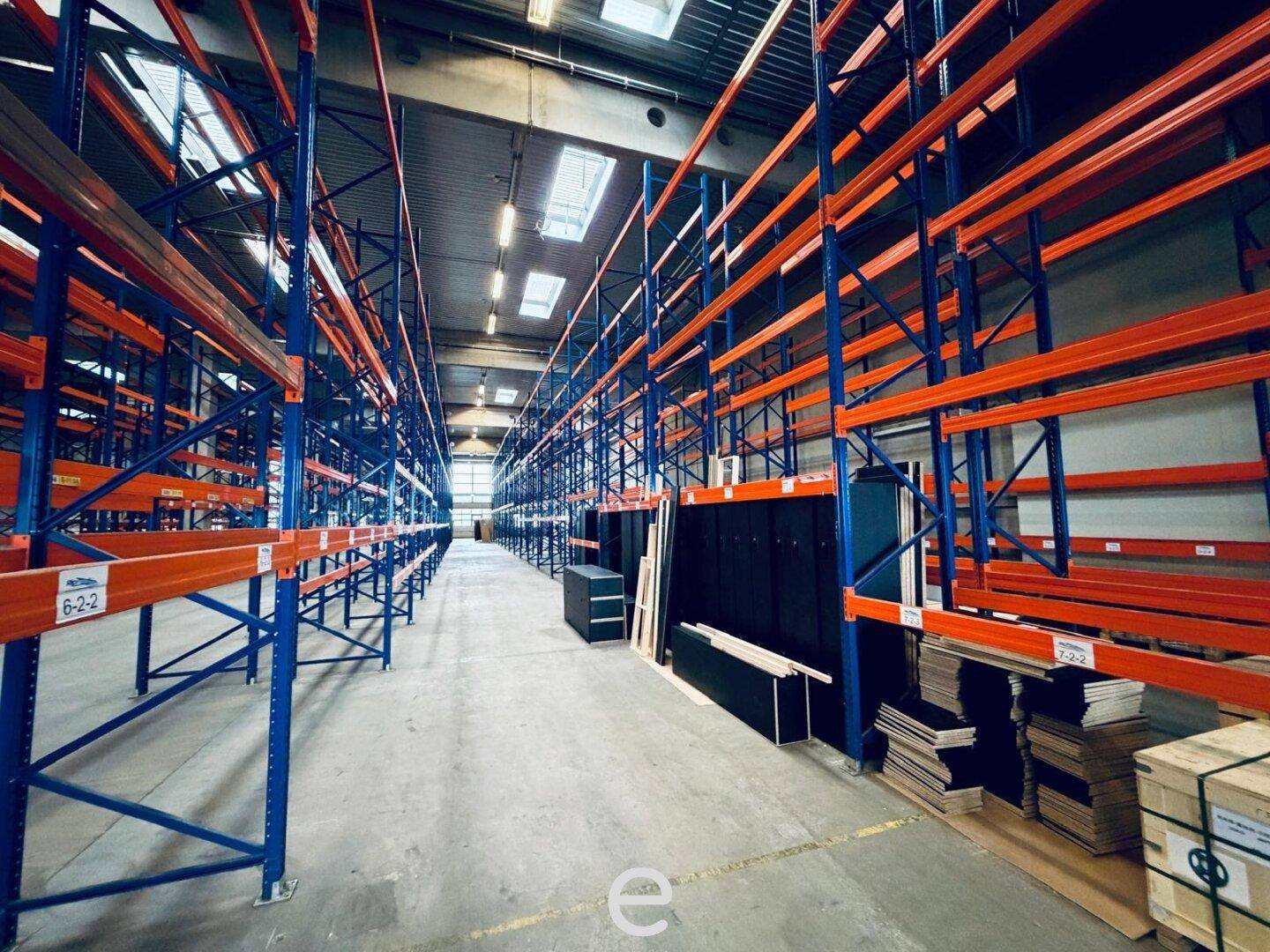 Langer Gang in einer Lagerhalle mit blauen und orangefarbenen Hochregalen und Betonboden.