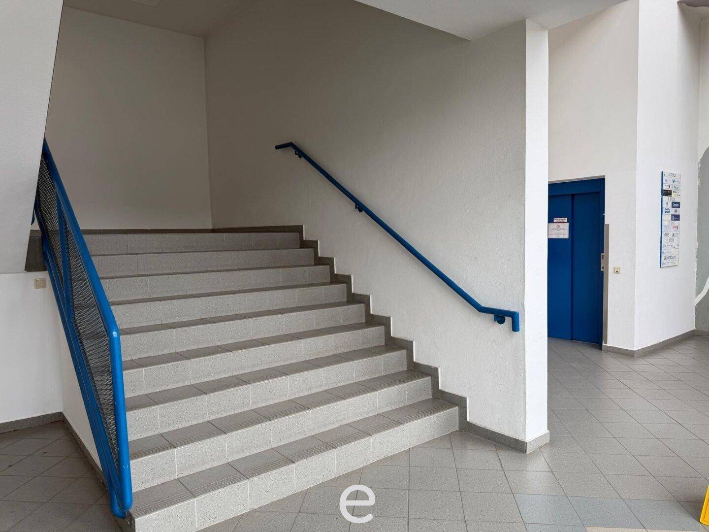 Treppenhaus mit gefliesten Stufen, blauem Geländer und Zugang zu einem Aufzug.