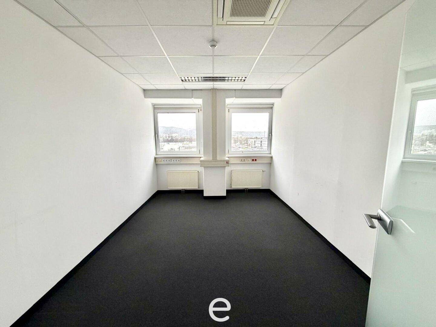 Kompaktes Büro mit dunklem Teppichboden, Klimaanlage und zwei Fenstern, bietet viel Tageslicht.
