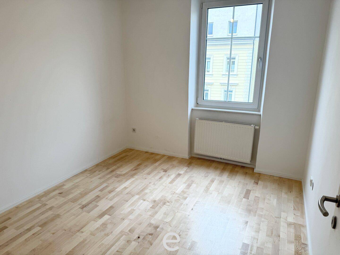 Leeres Zimmer mit hellem Parkettboden und Fenster zur Straßenseite.