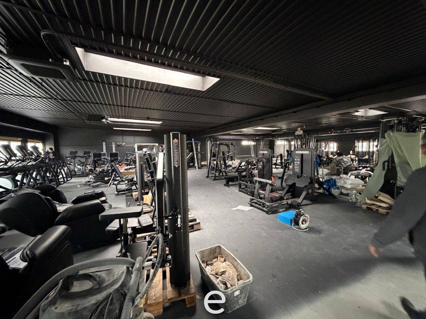 Großer Fitnessbereich mit verschiedenen Trainingsgeräten, dunklem Boden und hohen Decken.