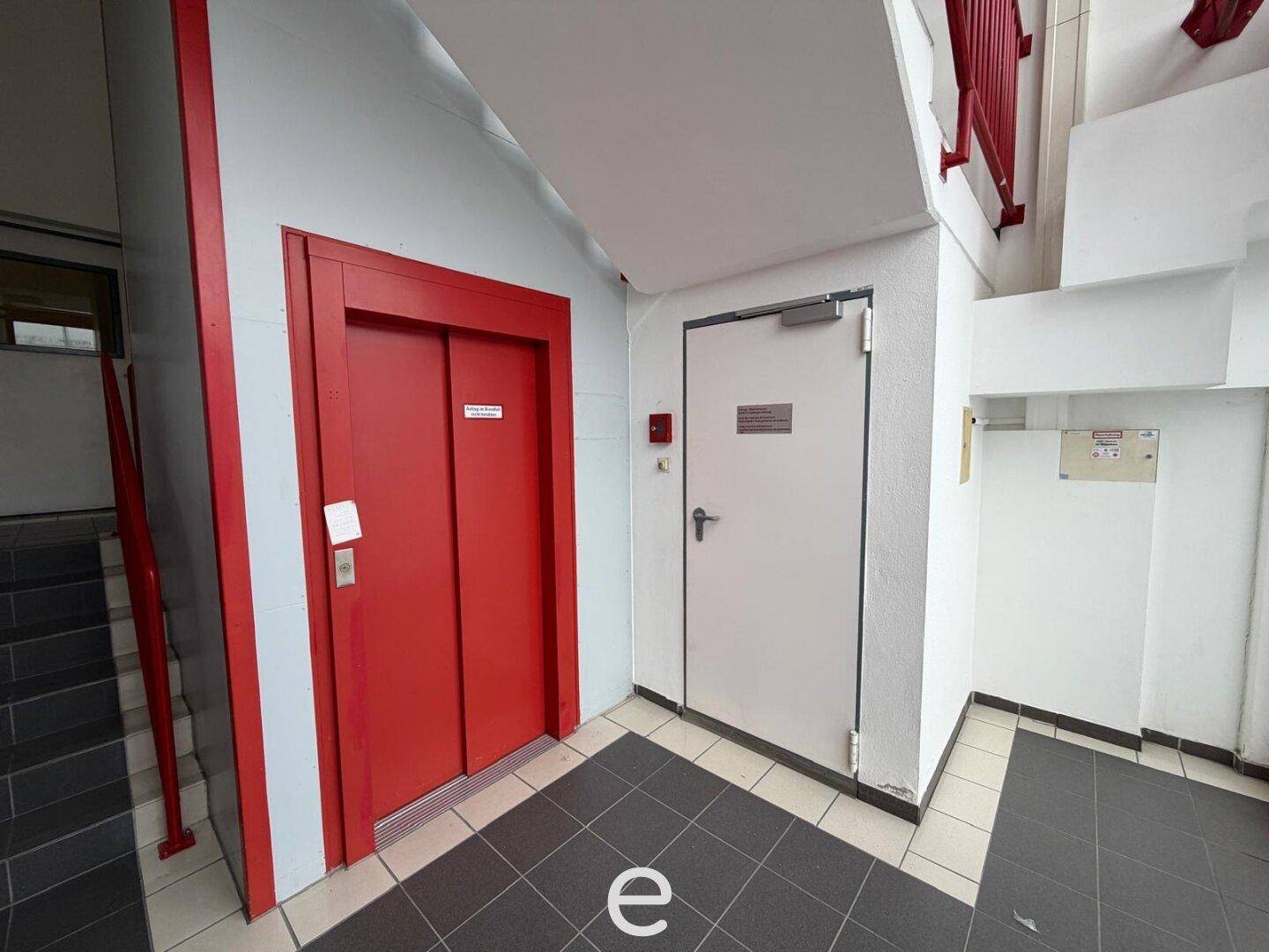 Eingangsbereich mit rotem Aufzug und Zugang zu den oberen Etagen, funktional und hell.