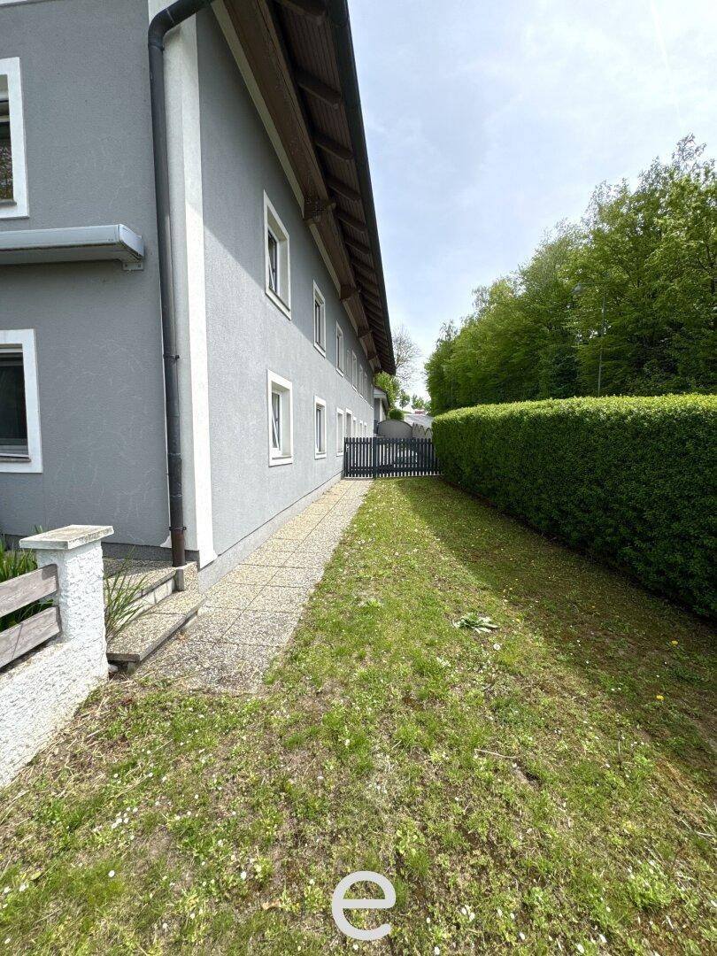 Gedeckter Weg neben dem Haus, der zu einer Rasenfläche und Hecke führt.