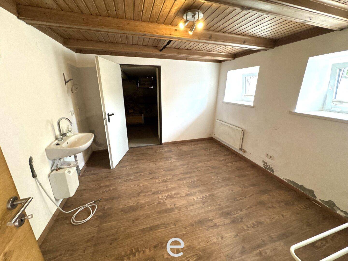 Einfacher Raum im Untergeschoss mit Waschbecken, Holzbalkendecke und Laminatboden.