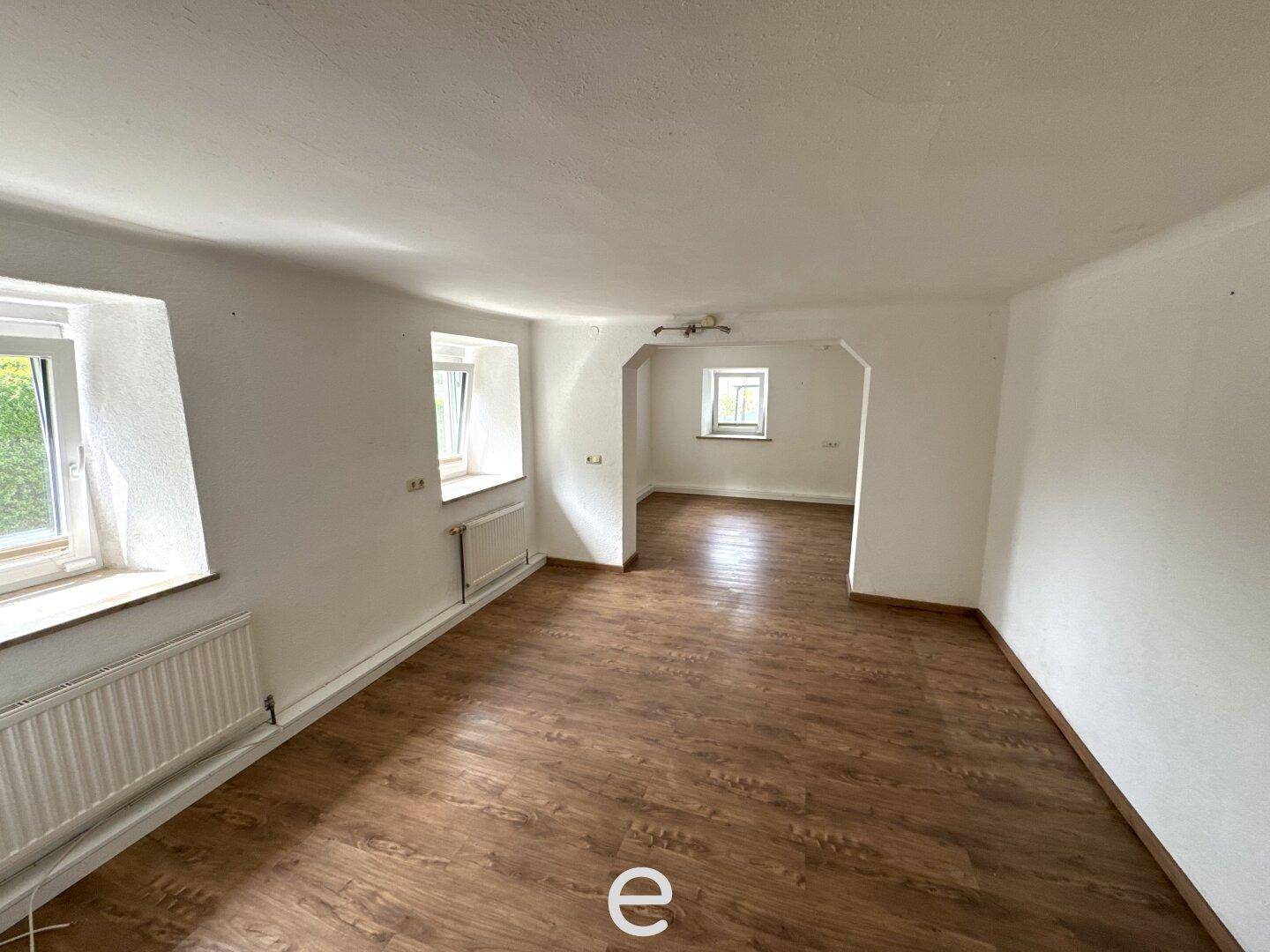 Geräumiges Wohnzimmer mit Laminatboden, weißen Wänden und mehreren Fenstern.