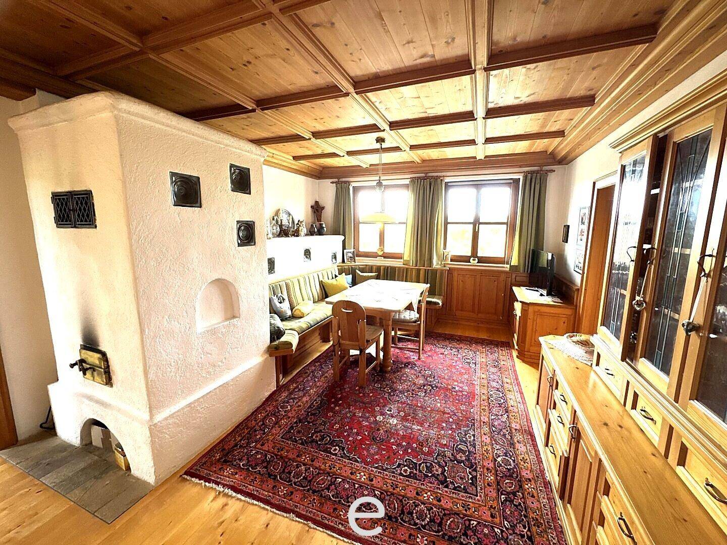 Gemütliches Wohnzimmer im alpinen Stil mit Kachelofen, Holzbalkendecke und traditionellem Teppich.