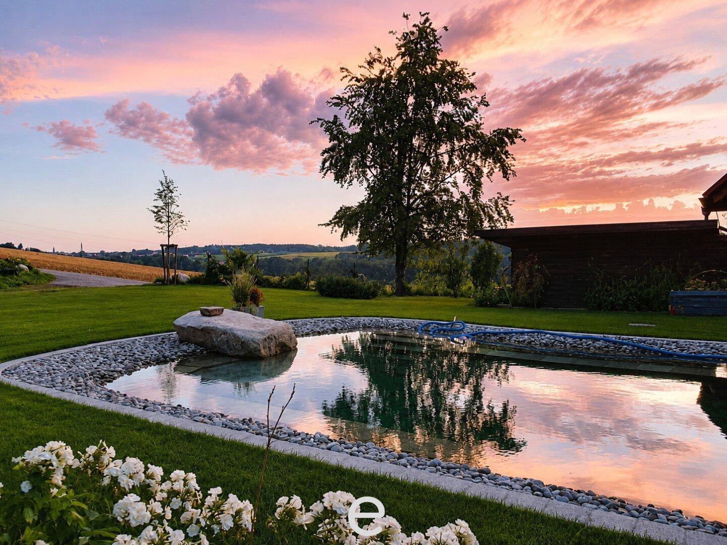 Naturnaher Poolbereich im Garten bei Sonnenuntergang mit blühenden Pflanzen und Baum.