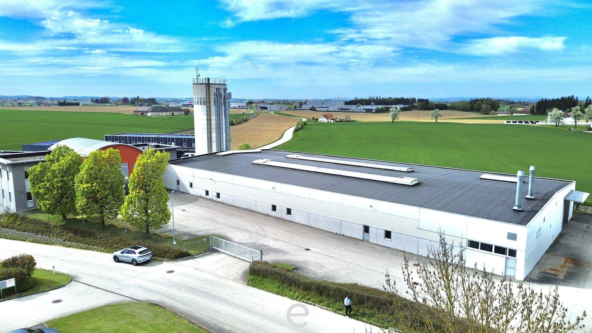 Luftaufnahme eines Industriegebäudes mit angrenzenden Feldern und einem Turm unter blauem Himmel.