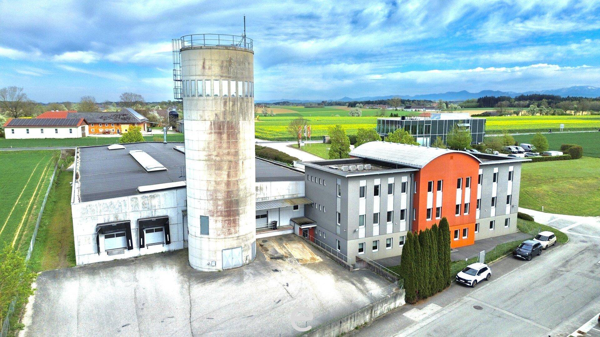 Industriekomplex mit einem hohen Silo und verschiedenen Gebäudeteilen, umgeben von ländlicher Landschaft.