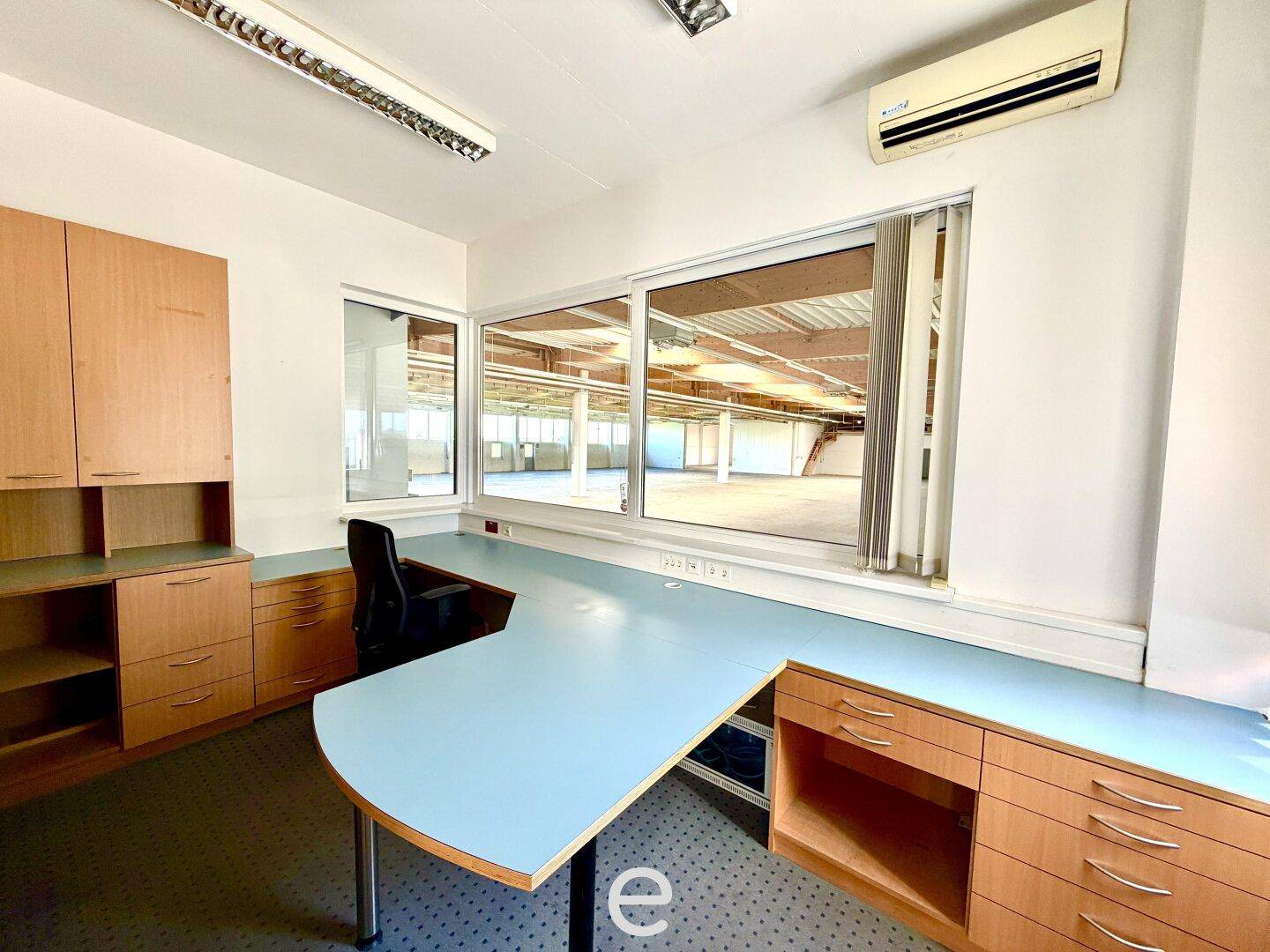 Einzelbüro mit großem Schreibtisch, Holzschränken und einem Fenster mit Blick in eine Halle.