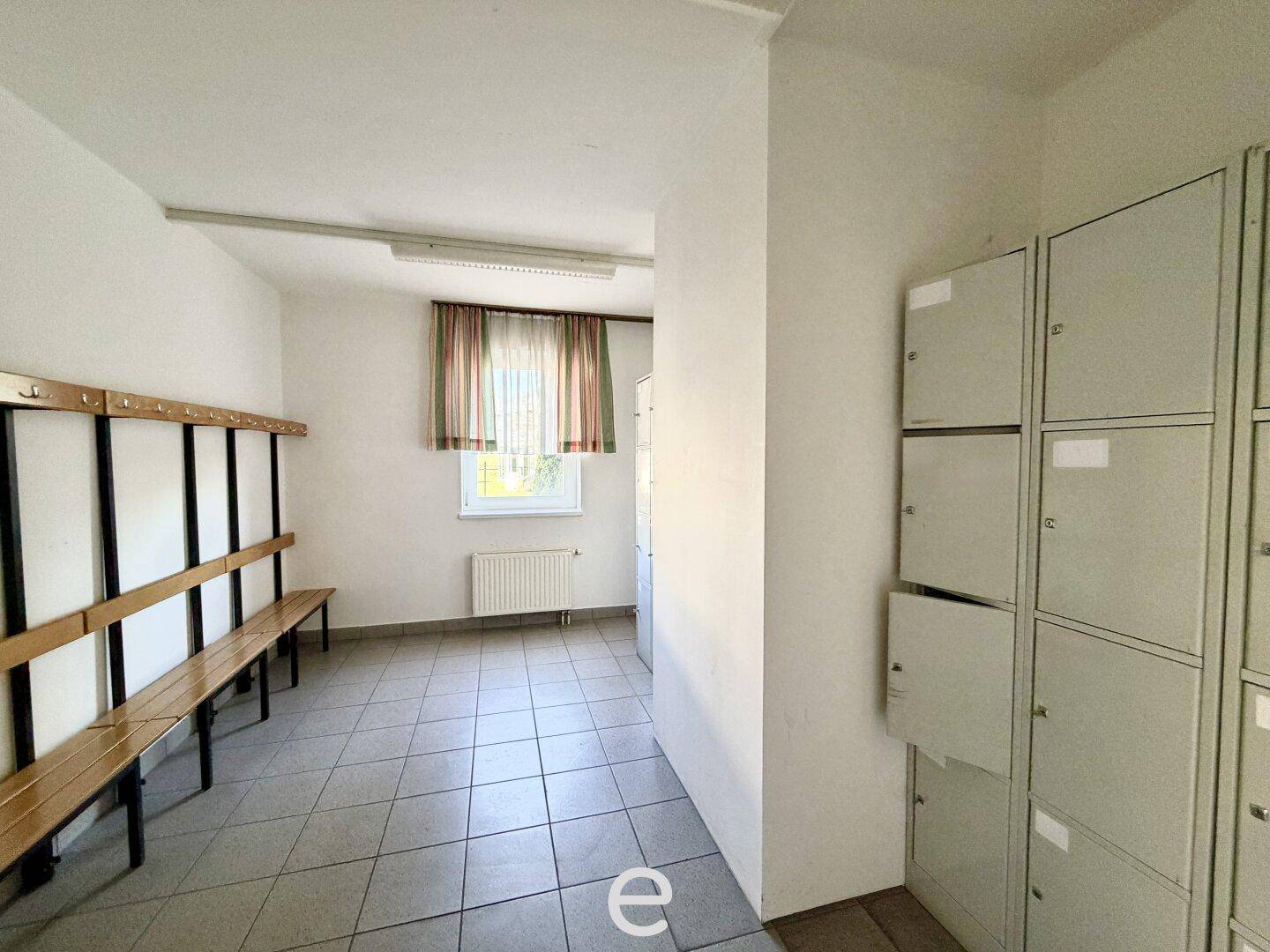 Umkleideraum mit Spinden, Sitzbänken und Fliesenboden, beleuchtet durch ein Fenster.