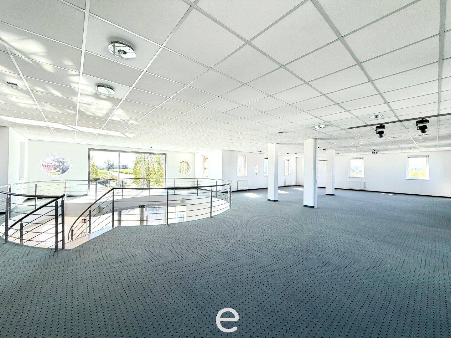 Großer, offener Bürobereich mit Teppichboden, Säulen und einer Galerie mit Glasgeländer.