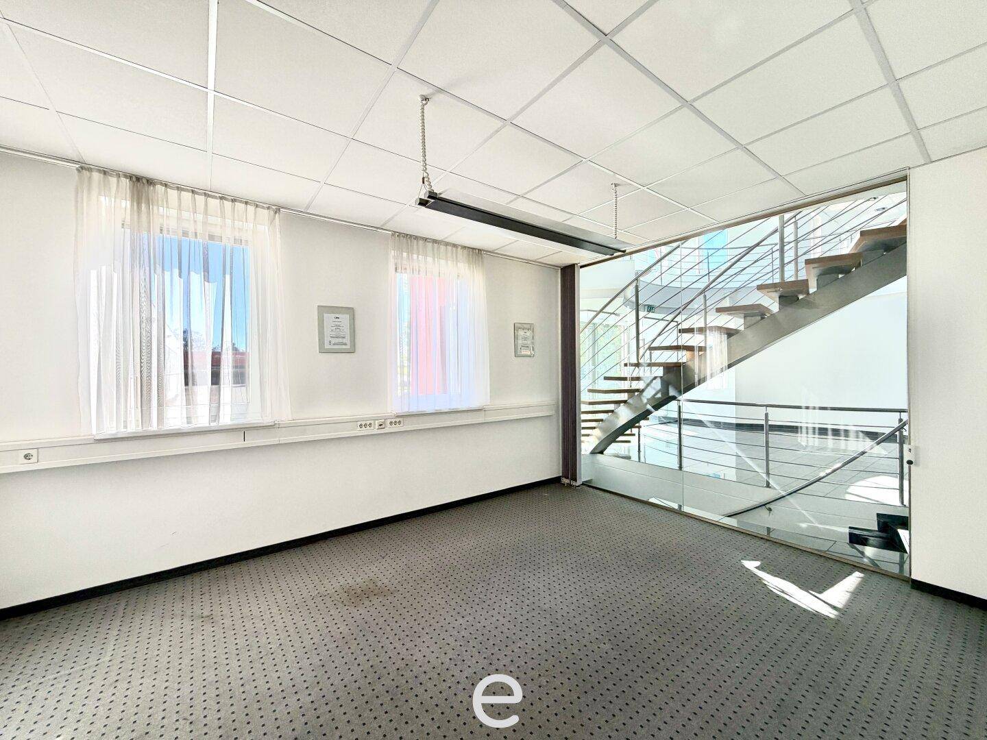 Heller Büroraum mit Teppichboden, großen Fenstern und einer modernen Treppe mit Glasgeländer.