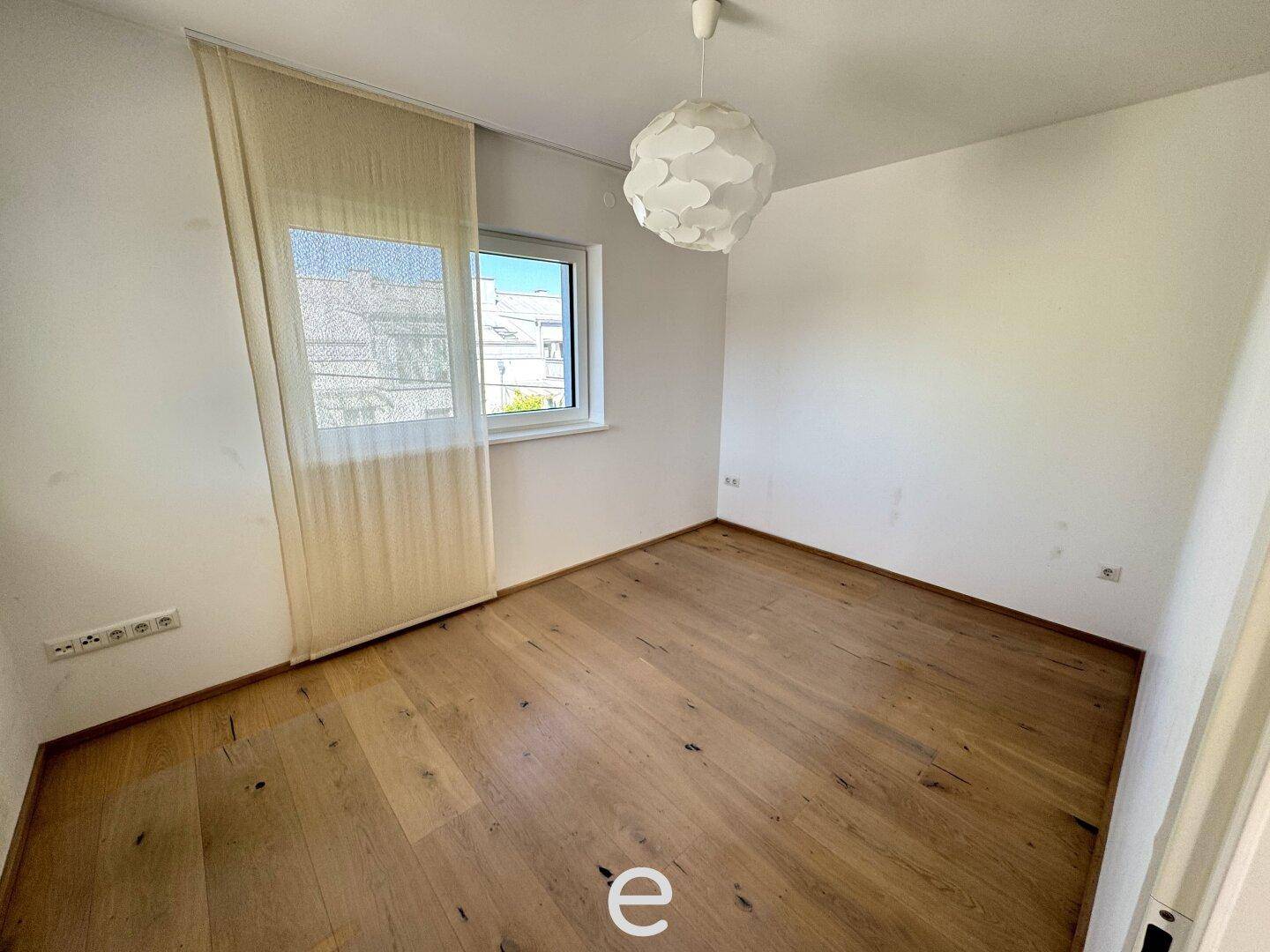 Gemütliches Schlafzimmer mit hellem Holzboden, Fenster und moderner Deckenleuchte.