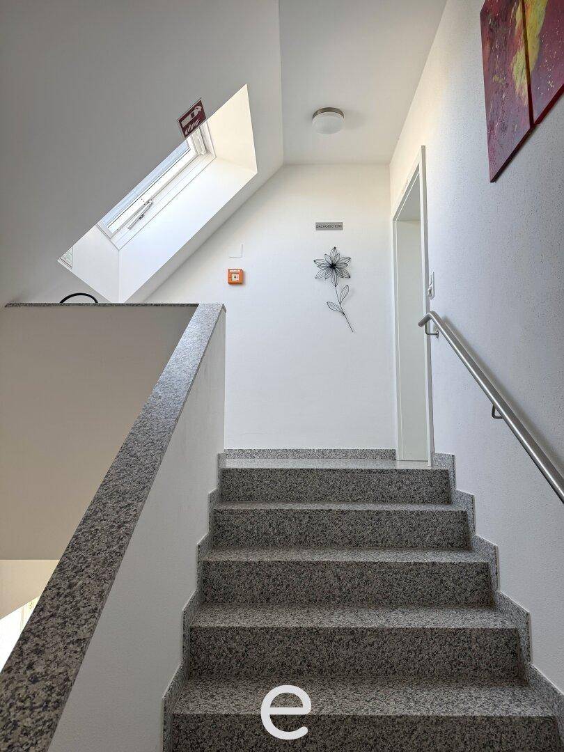 Helles Treppenhaus mit Granitstufen und Handlauf, beleuchtet durch ein Dachfenster.