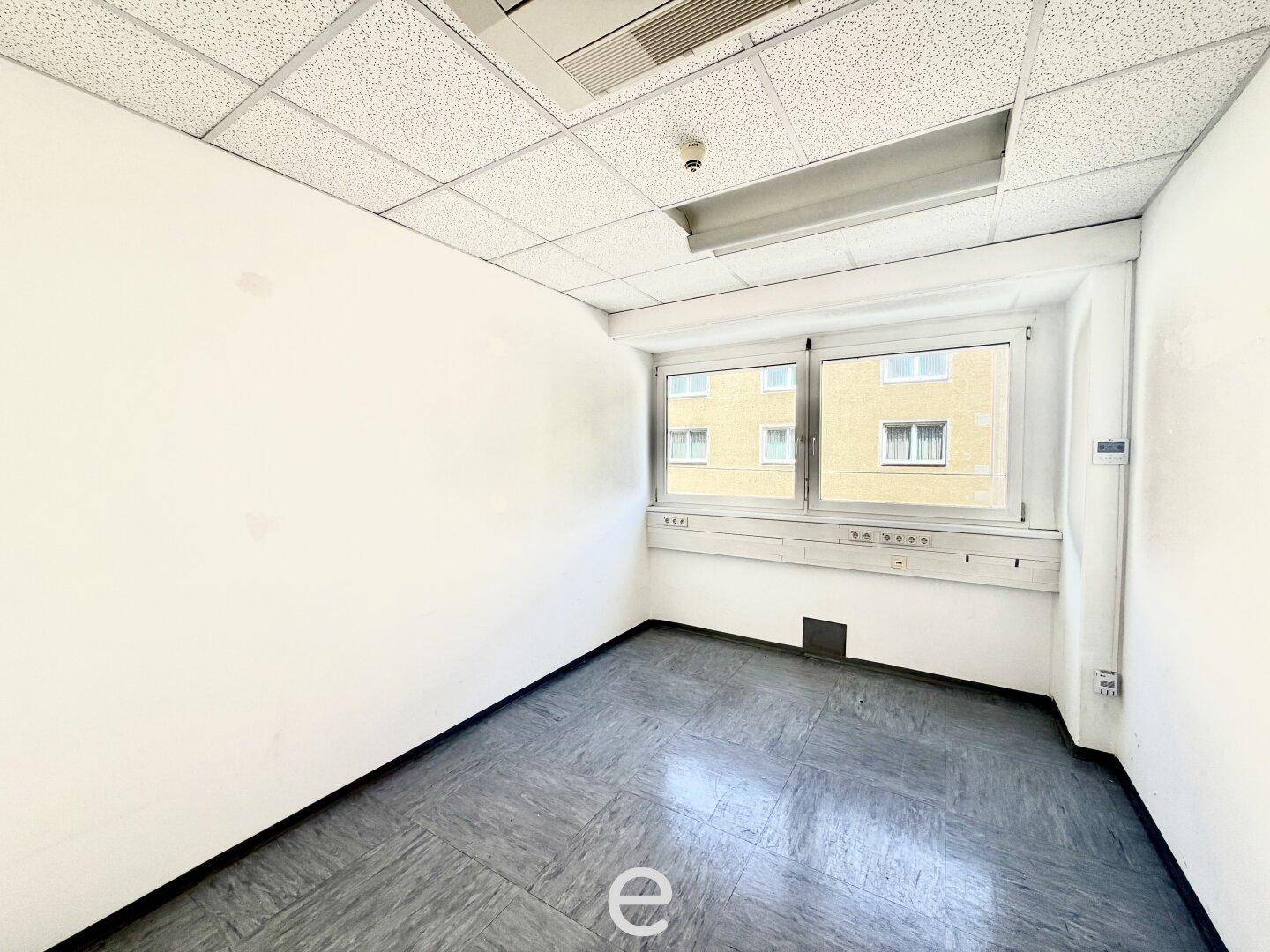 Kleineres, leeres Büro mit einem Fenster, abgehängter Decke und grauem PVC-Boden.