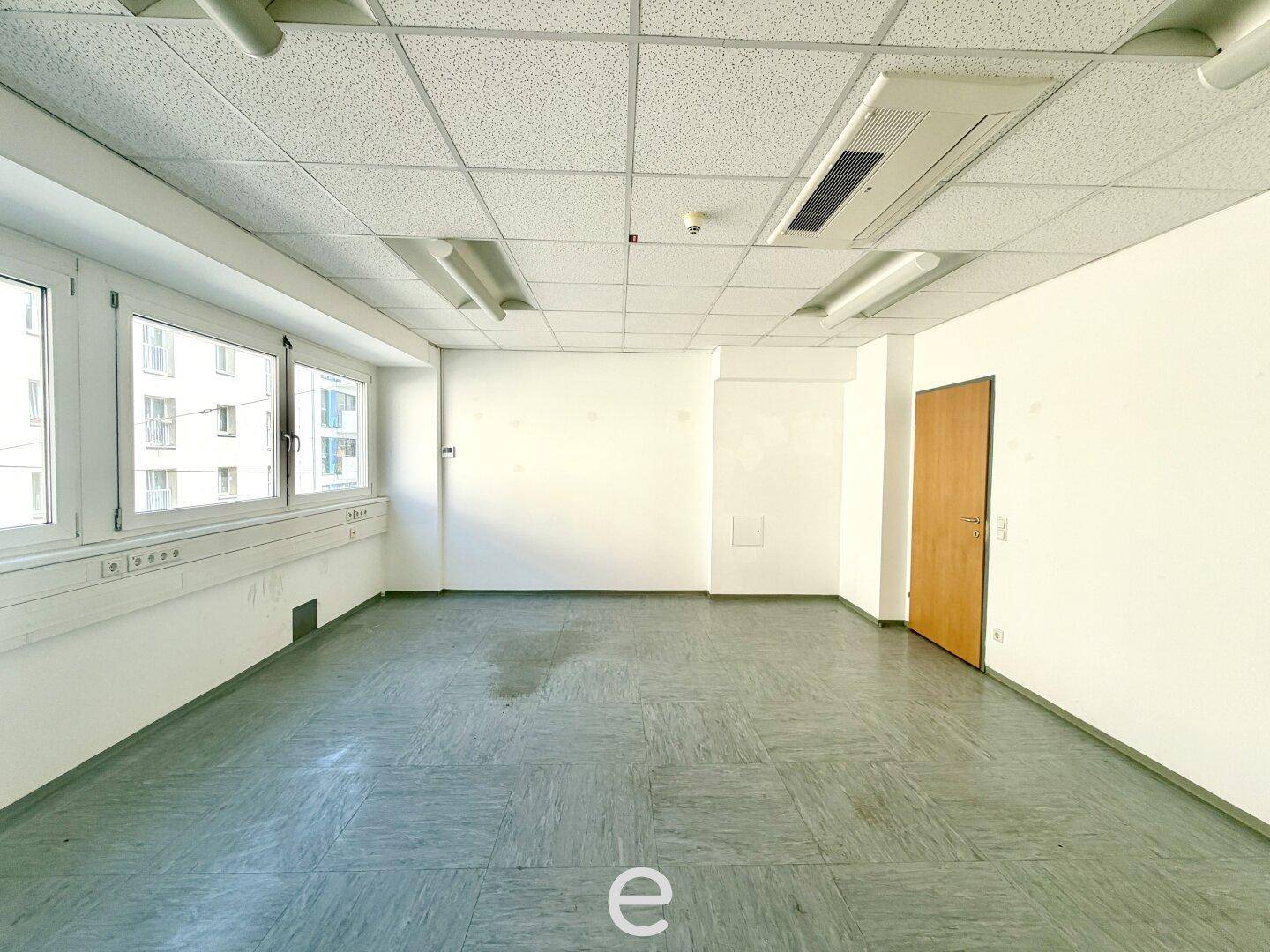 Geräumiges, leeres Büro mit vielen Fenstern, abgehängter Decke und grauem PVC-Boden.