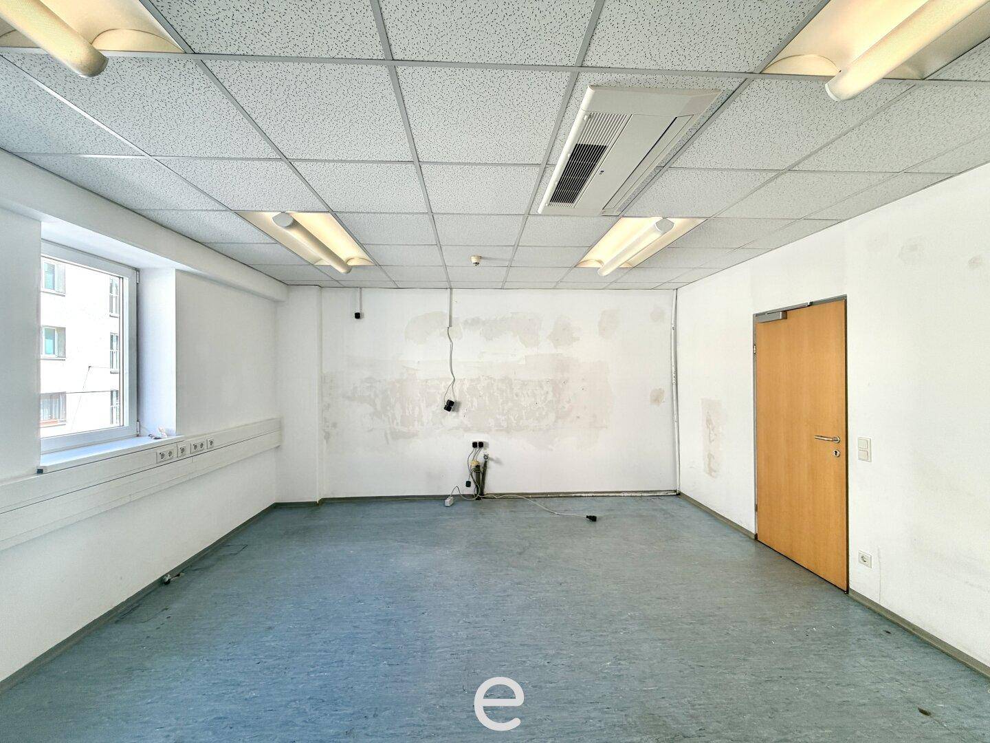 Leeres Büro mit einem Fenster, abgehängter Decke und blau-grauem Boden, renovierungsbedürftig.