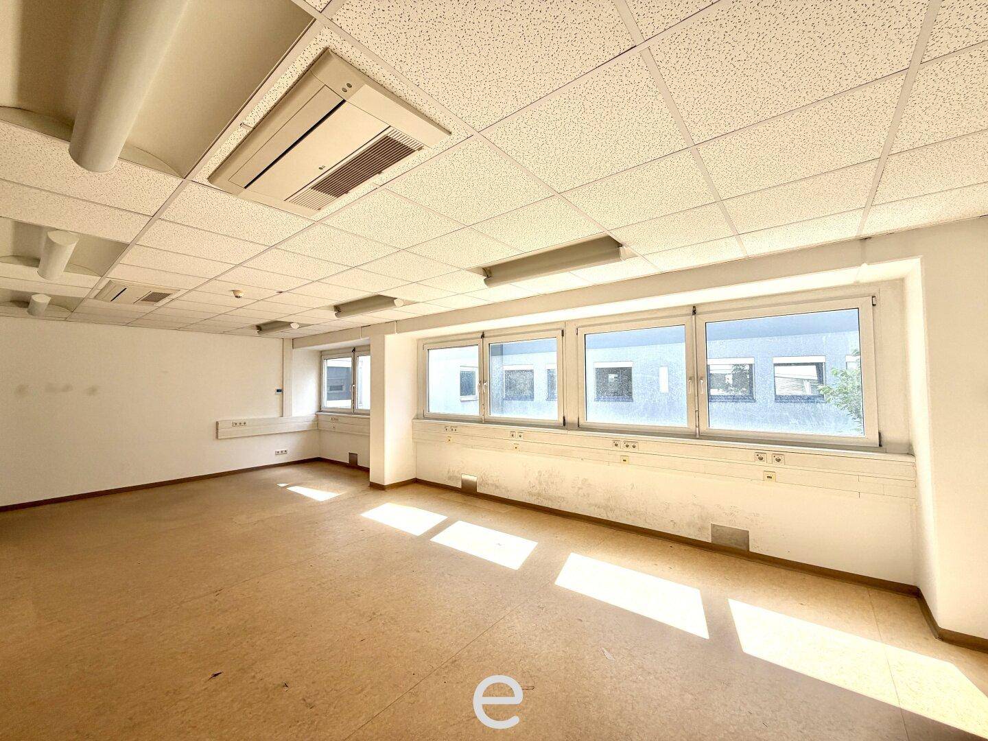 Geräumiges, leeres Büro mit großen Fenstern und hellem Linoleumboden, viel Tageslicht.