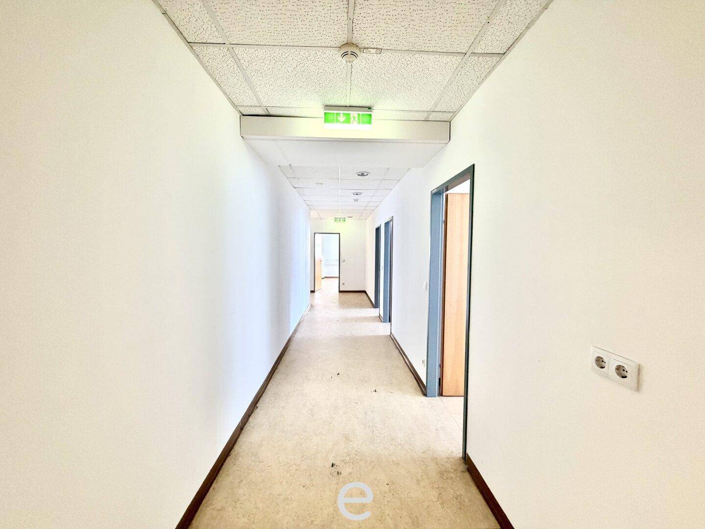 Langer Büroflur mit Linoleumboden und Notausgangsbeschilderung, gut beleuchtet.