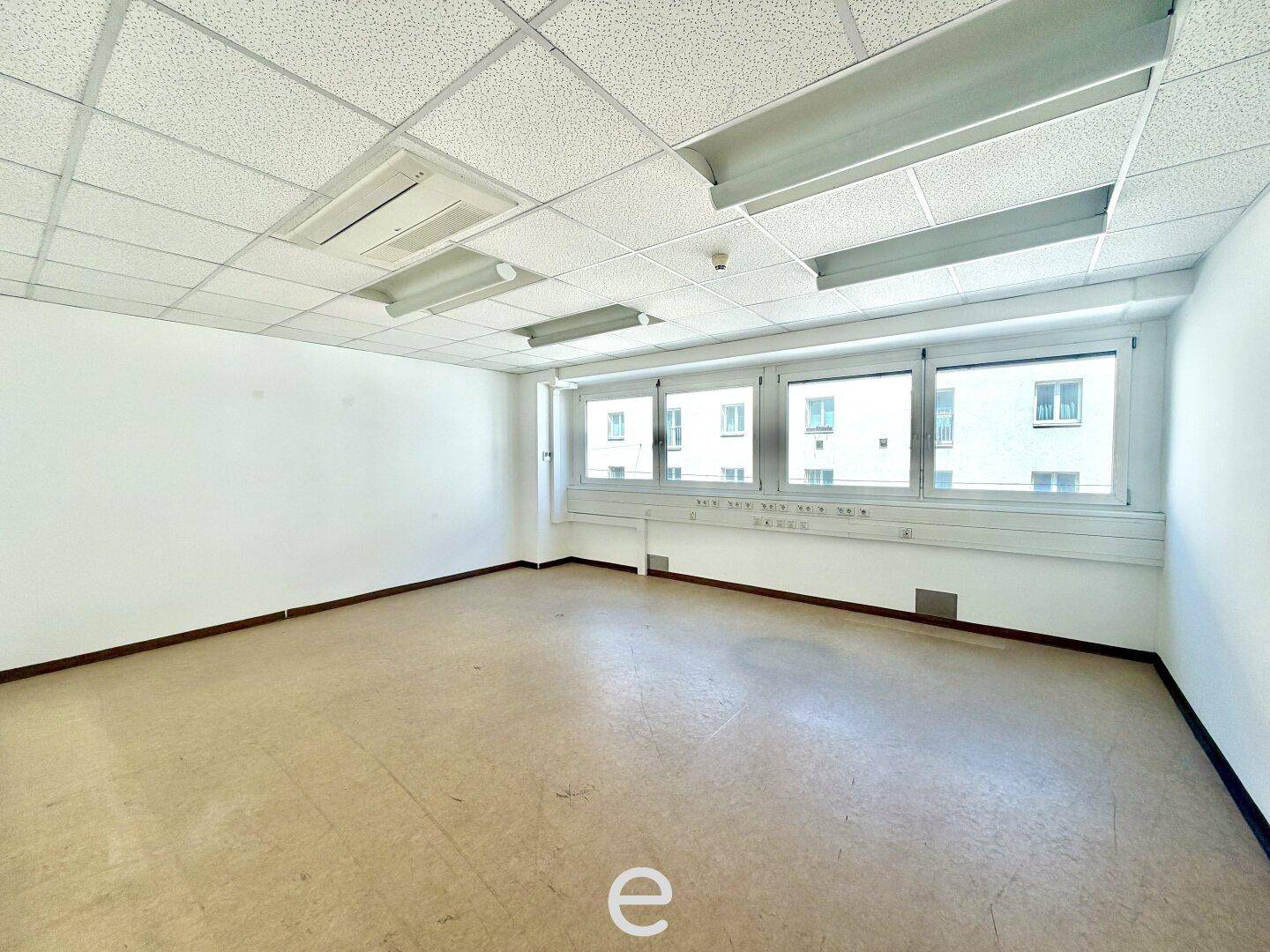 Geräumiges, leeres Büro mit großen Fenstern und hellem Linoleumboden, viel Tageslicht.
