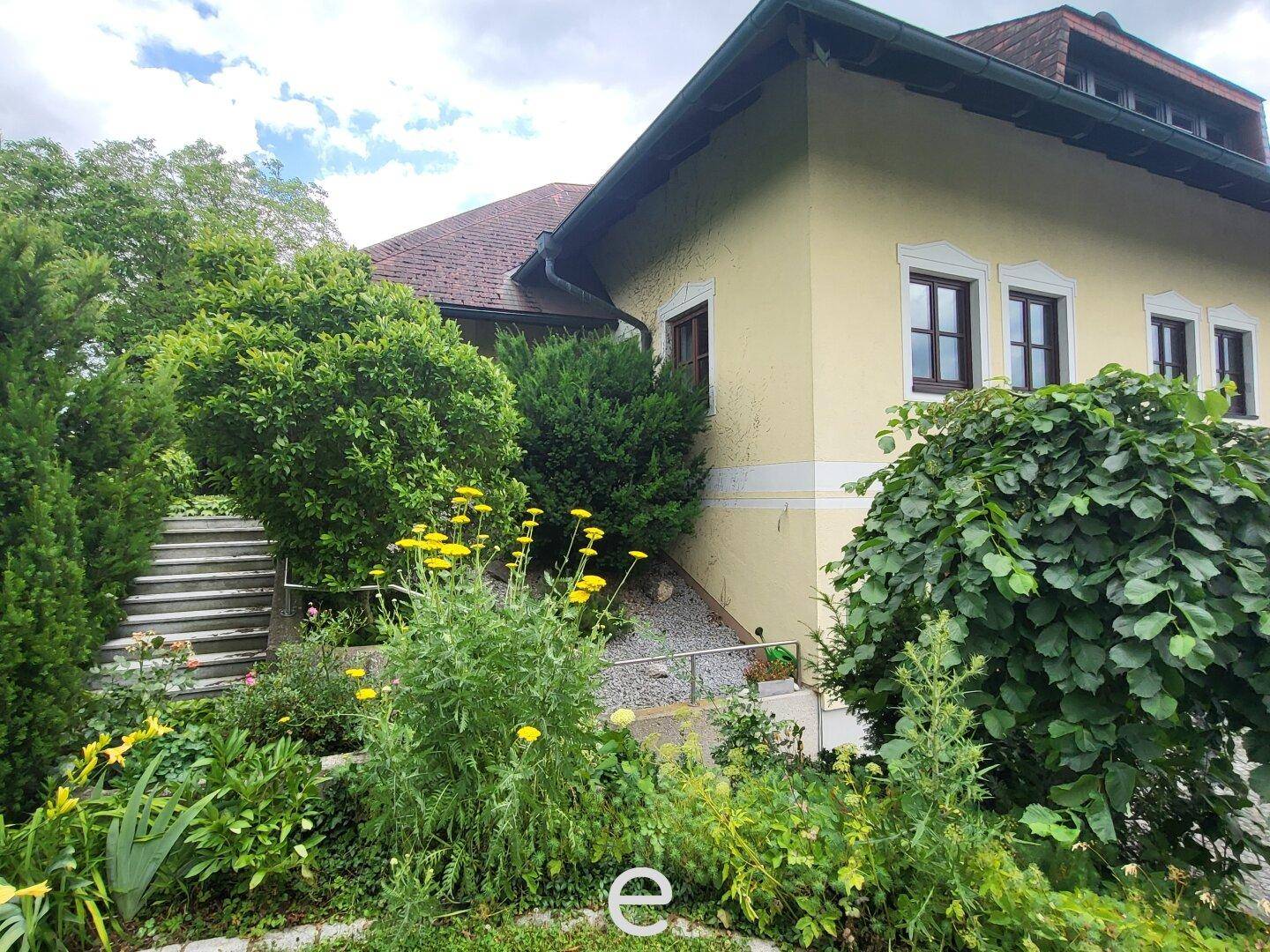 Seitenansicht des Hauses mit blühenden Pflanzen, Sträuchern und einer Steintreppe.
