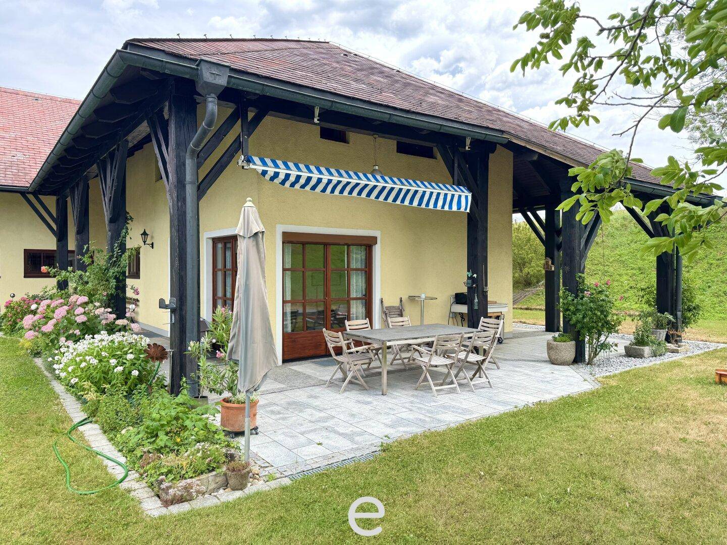 Überdachte Terrasse mit großem Esstisch, Markise und blühenden Pflanzen im Garten.