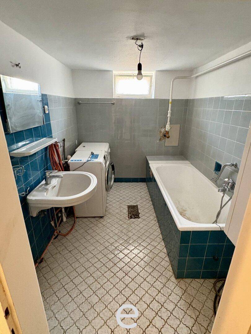 Älteres Badezimmer mit Badewanne, Waschbecken und Waschmaschine, gefliest in Blau- und Grautönen.