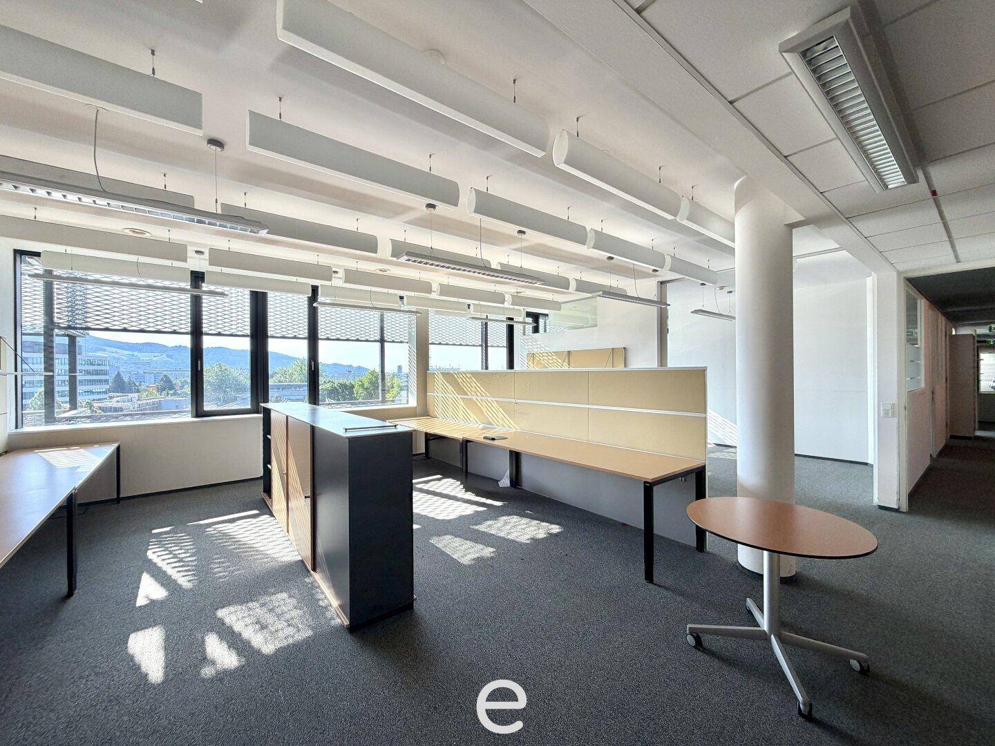 Offener Bürobereich mit Schreibtischen, Trennwänden, Teppichboden und großen Fenstern mit Weitblick.