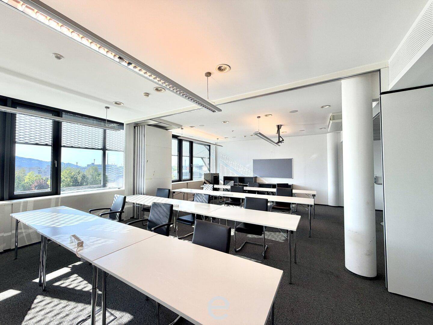Konferenz- oder Schulungsraum mit mehreren Tischen und Stühlen, Teppichboden und großen Fenstern für Tageslicht.