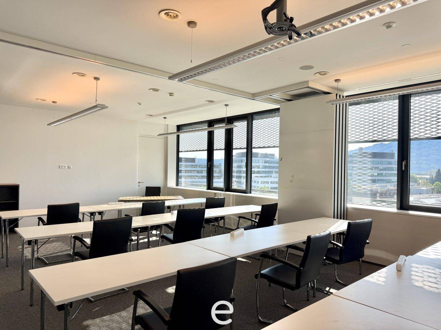 Konferenz- oder Schulungsraum mit mehreren Tischen und Stühlen, Teppichboden und großen Fenstern für Tageslicht.