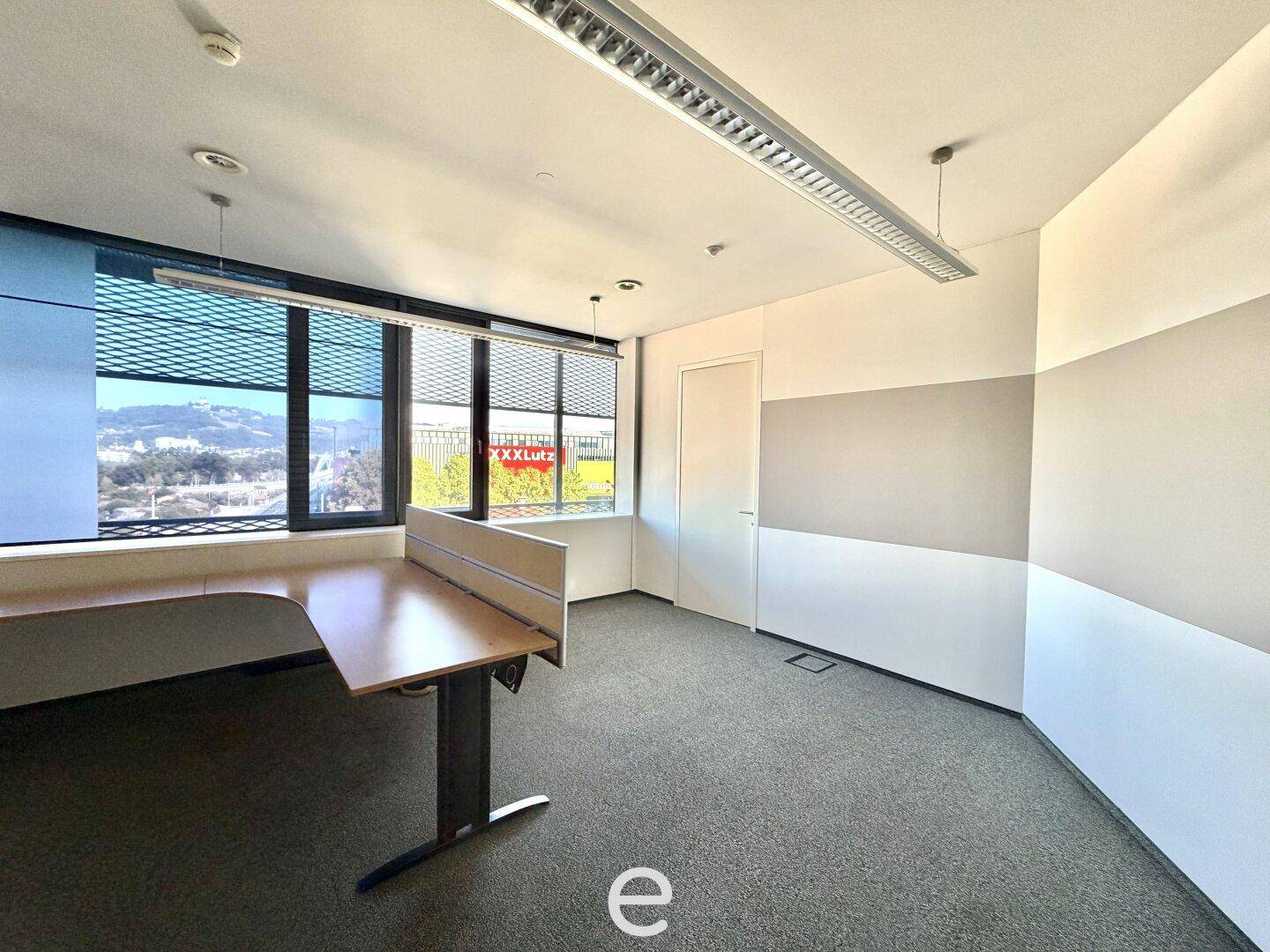 Büroraum mit Schreibtisch, Trennwand, Teppichboden und großen Fenstern, die viel Tageslicht hereinlassen.