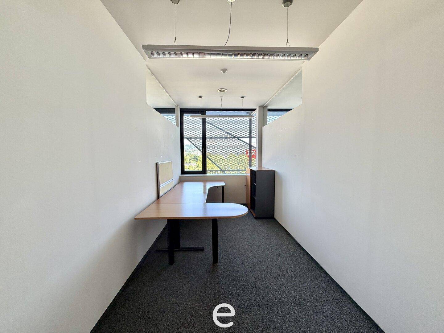 Kleiner Büroraum mit Schreibtisch, Teppichboden und einem Fenster, das Tageslicht spendet.