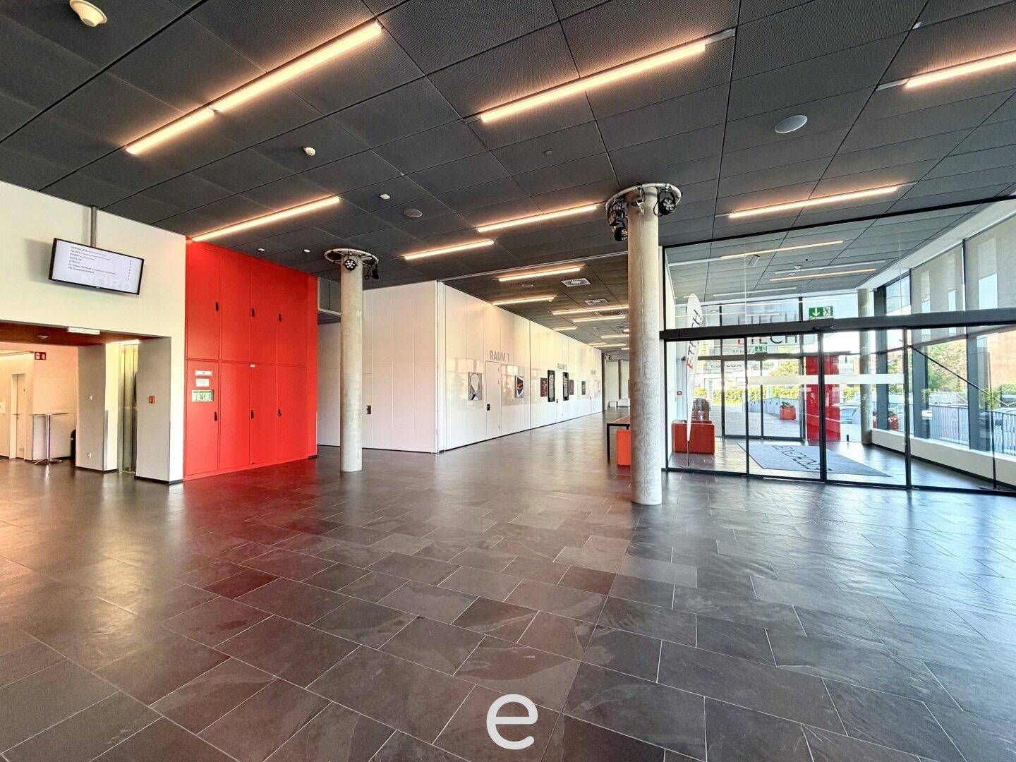 Geräumige, moderne Lobby mit dunklem Fliesenboden, roten Akzentwänden und großen Glasfronten am Eingangsbereich.