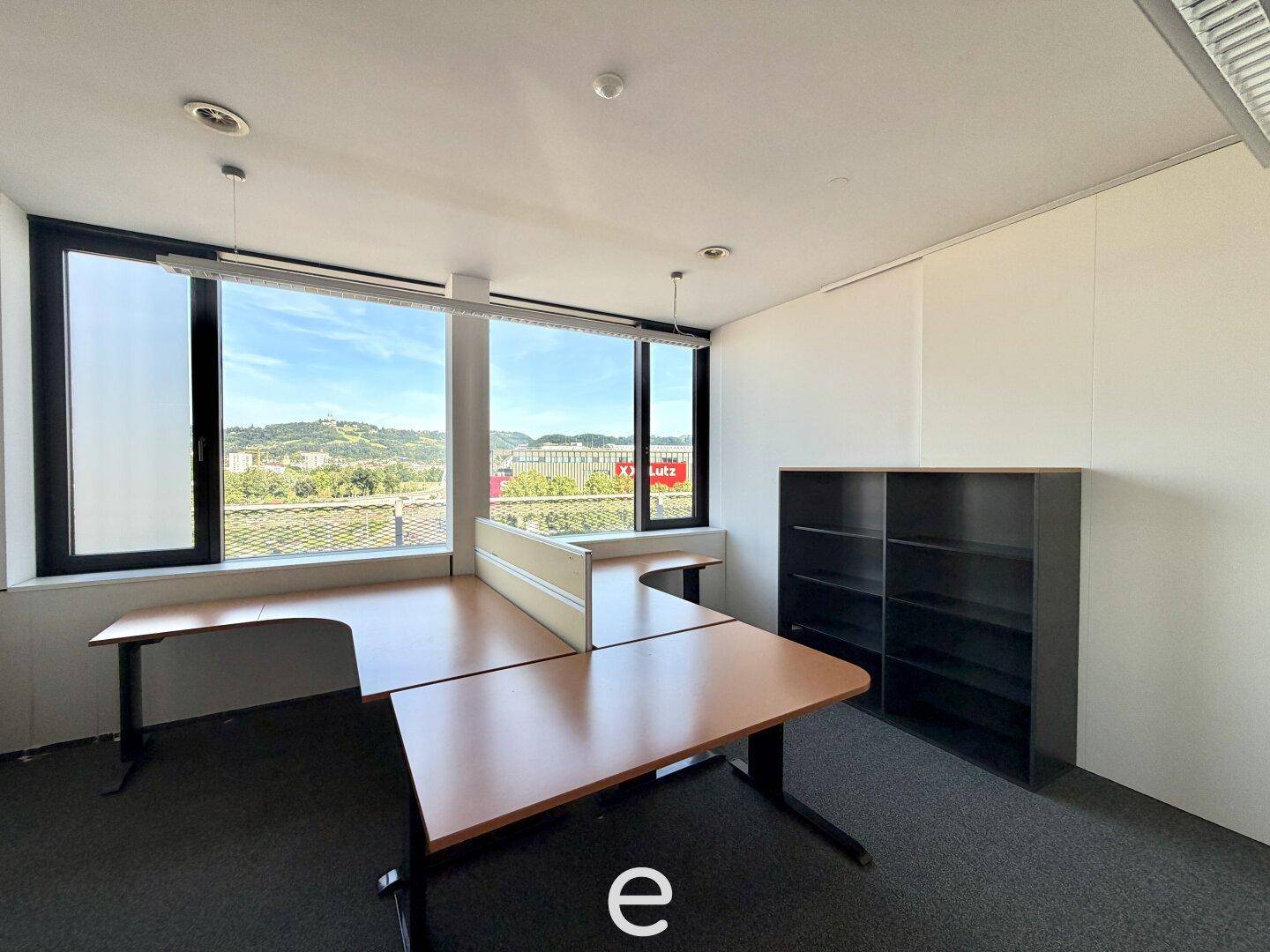 Offener Bürobereich mit mehreren Schreibtischen, Regalen, Teppichboden und großen Fenstern mit Ausblick.