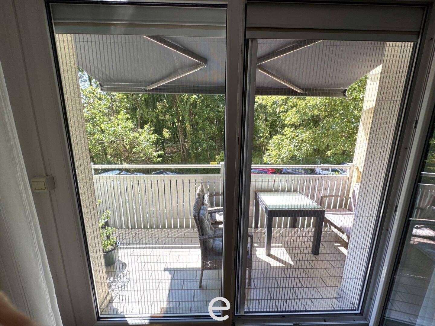 Blick vom Innenraum durch eine Glastür auf den möblierten Balkon mit grüner Umgebung.