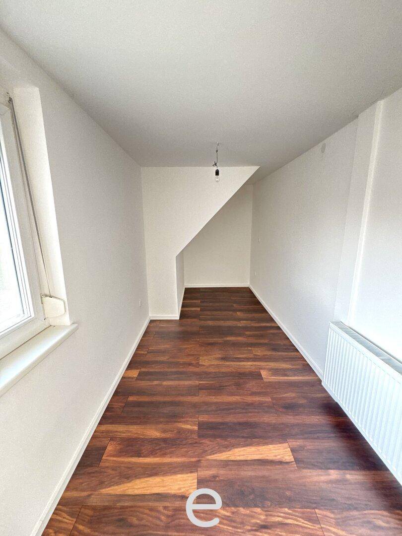 Schmaler Raum mit dunklem Laminatboden, Fenster und einer Dachschräge, ideal als Arbeitsbereich.